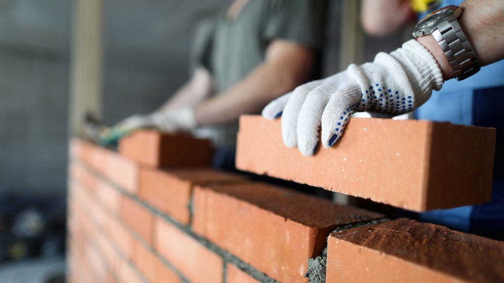Two workers making red brick wall at construction site close-up